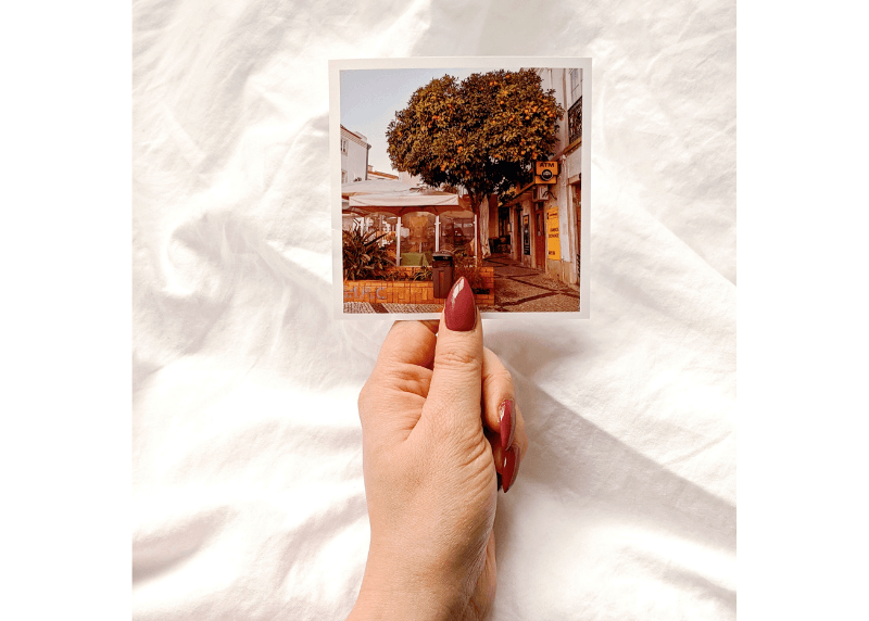 A woman's hand holds a picture of an orange tree in old town lagos portugal, over a white blanket.