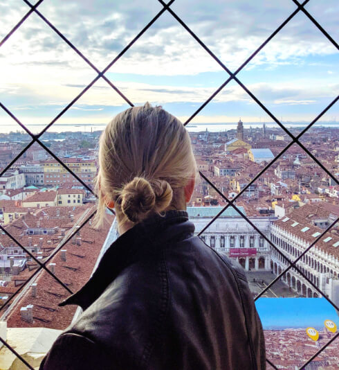 A woman with blonde hair twisted tightly into a bun, looks through a diamond shaped fence onto St. Mark's square and the roofs of Venice below.
