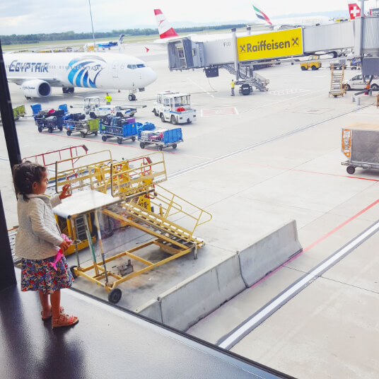A small girl stands on an arrivals ramp, looking through the glass onto the planes and airport equipment below. Vienna, Austria.