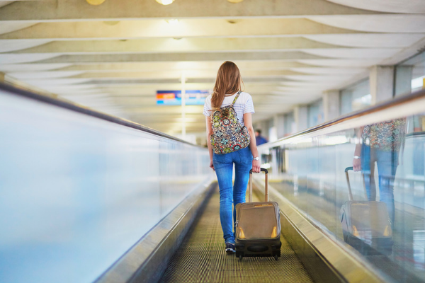 A woman with long brown hair walks on a moving walkway in the airport, wearing a backpack and pulling a small suitcase.