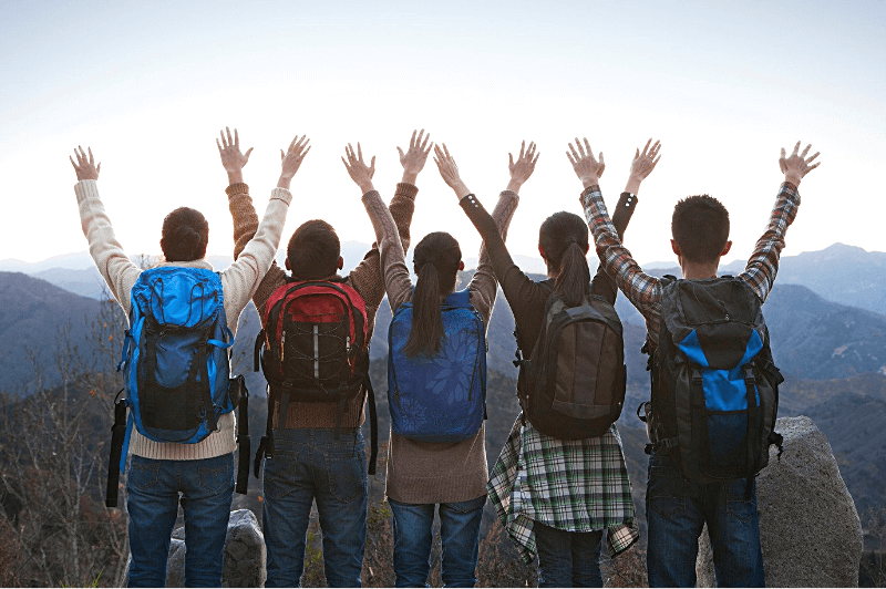 a set of five friends in backpacks raise their arms over a mountain view