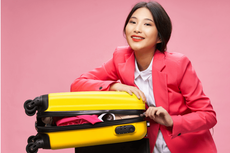a woman in a pink blazer smiles as she squishes her yellow suitcase closed