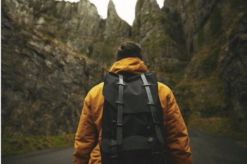 A man in a mustard coat hikes through the forest with a Little America backpack