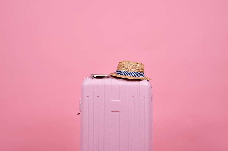 A pale pink suitcase in front of a pink wall with a straw hat resting on top of it.
