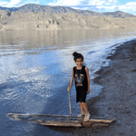 A girl stands on a beach with one foot on a piece of driftwood.