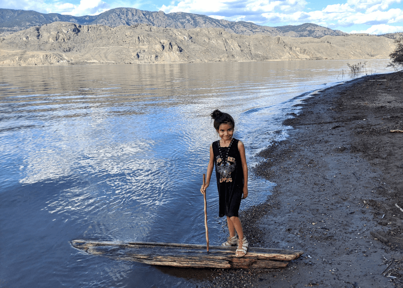 A girl stands on a beach with one foot on a piece of driftwood.