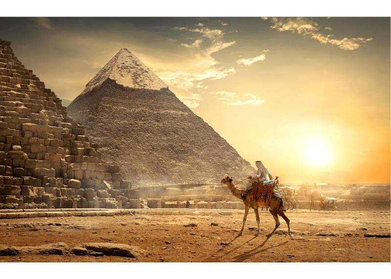 A man rides a camel past the pyramids when the sun is low in the sky