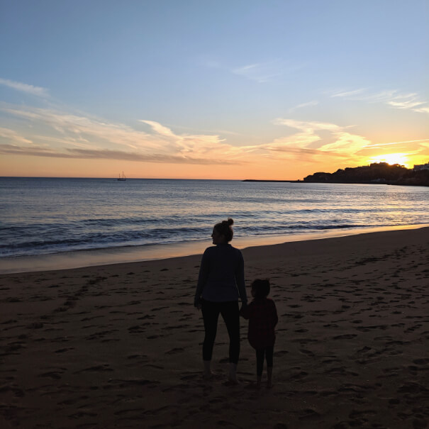 A woman and little daughter holding hands on the beach, silhouetted by the setting sun over the ocean in Albufeira Portugal.