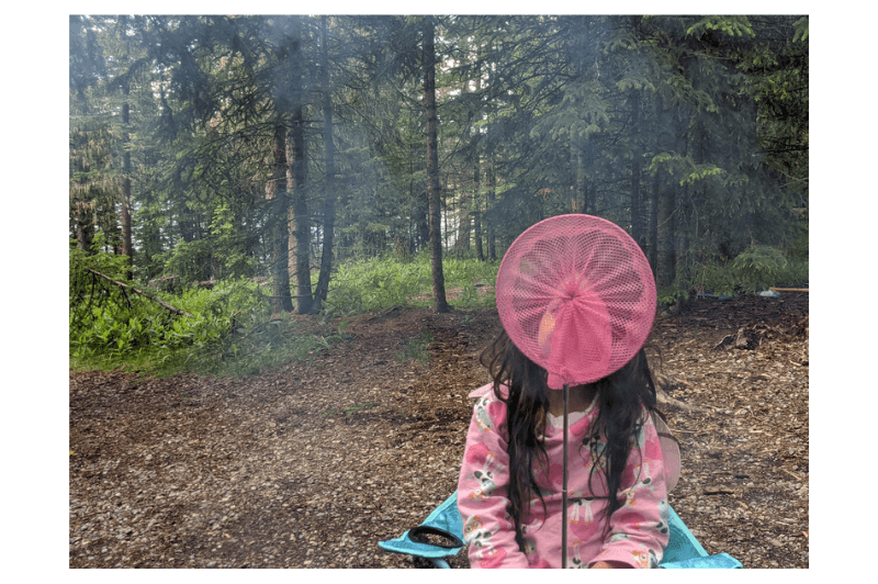 A young girl holds a pink butterfly net in front of her face as she sits in the forest camping