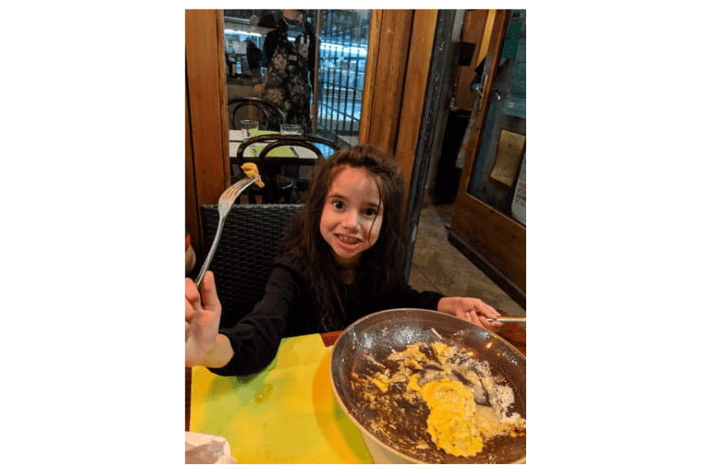 A girl smiles at the camera and holds up a piece of ravioli on a fork