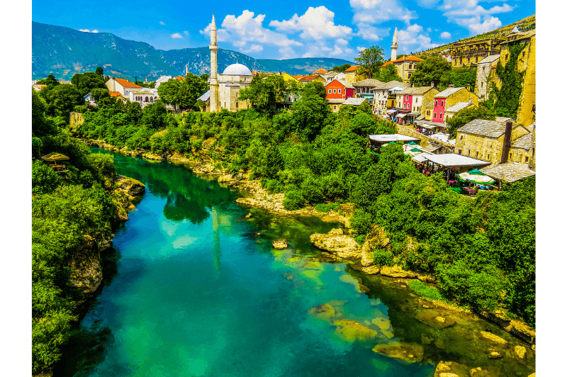A colourful photo of a town in Bosnia and Herzegovina. A colourful turkish inspired village beside a blue-green river with green shrubs and trees on either side.