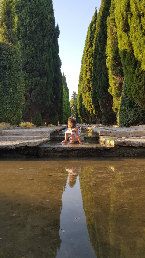 A young girl trails her hand into a manmade stream as she sits on a stepping stone. Tall cedars rise in the distance on either side of the water feature at Balchik Palace in Bulgaria
