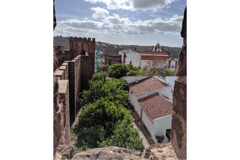 A view through a brick window in the ruins of a dark red castle in Silves. The white village sits below with red clay roofs.