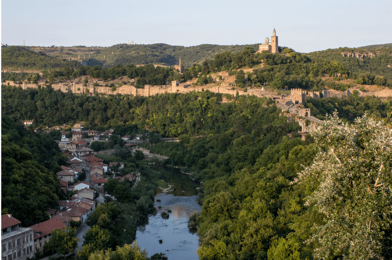 A quiet river runs through the village of Veliko Tarnovo. The village is on the left and the right bank is covered in shrubs and trees, rising to meet the fortress walls of Tsaravets