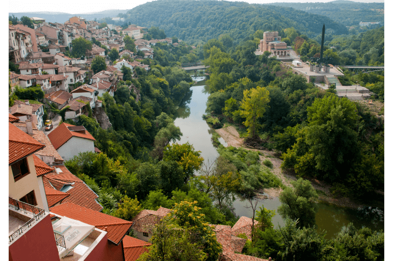 A quiet river runs through the village of Veliko Tarnovo. The village is on the left and the right bank is covered in shrubs and trees, rising to meet the fortress walls of Tsaravets