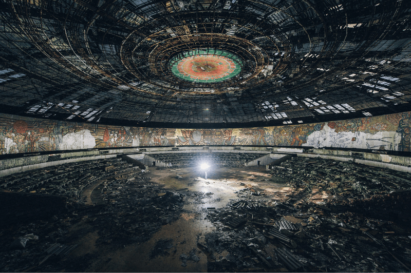 A view inside Buzludzha Monument. The stained glass hammer and sickle is in the centre of the circular building on the ceiling. There is broken glass everywhere and daylight shines through the dark.