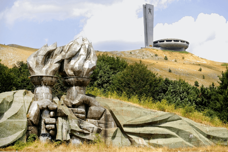 A stone sculpture of two hands holding torches up to each other in front of a hill with a futuristic looking round concrete building at the top