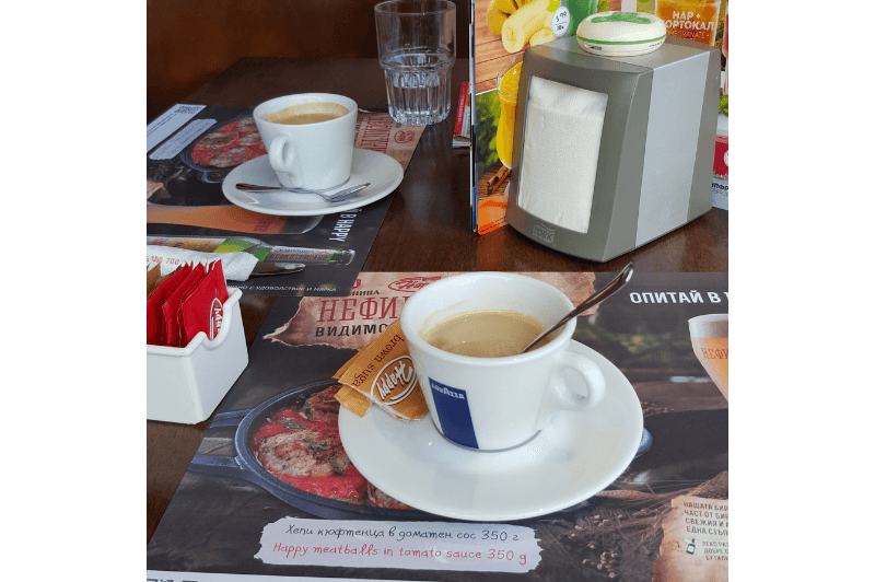 Two espresso cups and saucers on paper placemats with cyrillic writing on them, sugar packets sit on the table to the left, and a retro napkin holder rests on the right.