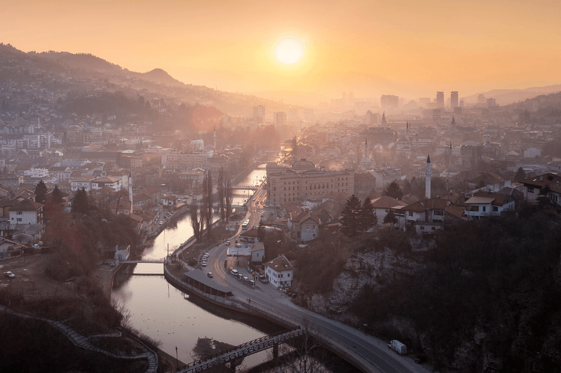 The city of Sarajevo on a winter's morning. The sun is low in the sky behind a haze of frost and smoke. The rive shines in the middle of the city which is built up around it. A light blanket of snow rests on the city