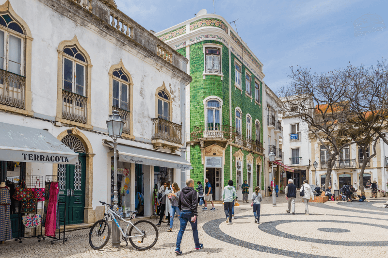 Adorable buildings in the centre of Lagos Portugal on one side of a square, the foreground is a white stucco two storey house with juliette balconies on the second floor and shops on the first. A man walks past a parked bicycle. A green tile 3 storey building sits across the street. Two trees sit centre square and the cobbles have a black and white circular pattern.