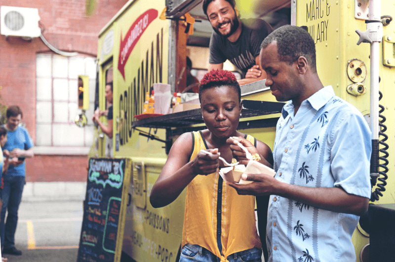 A couple try food from a yellow truck as the truck operator smiles behind them.