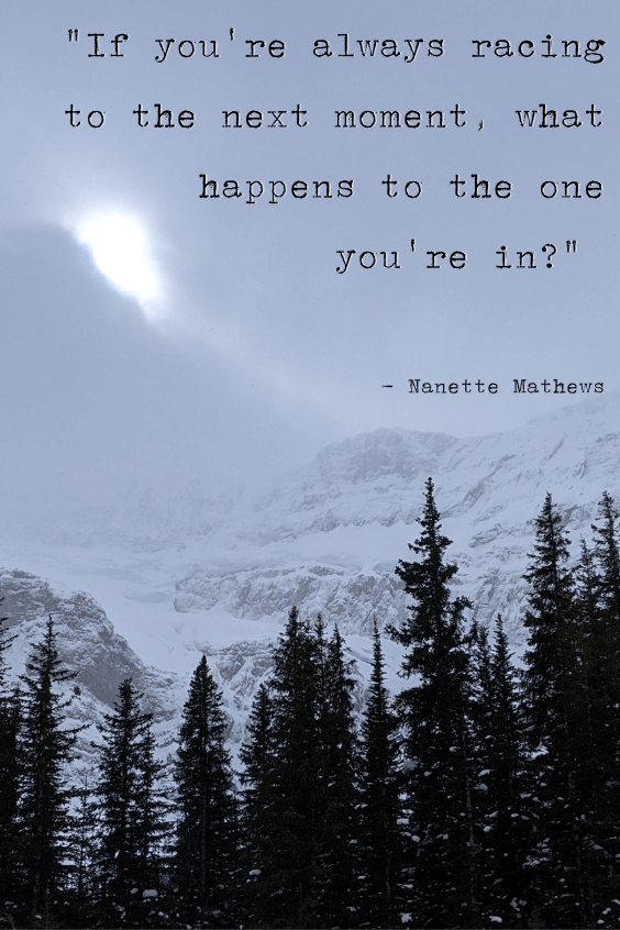 Snow covered Rocky Mountains with a dark forest in front of them. The sun hides, glowing white behind some white clouds. Quote on top reads "If you're always racing to the next moment, what happens to the one you're in?" - Nanette Mathews