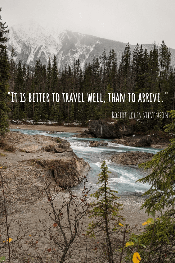 A light blue green river runs through some rocks with a forest around it. Rocky Mountains hide in some low cloud in the background. Quote reads "It is better to travel well than to arrive." - Robert Louis Stevenson