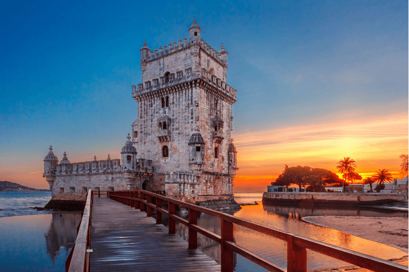 Belem tower in Lisbon Portugal at sunset. The orange glow of the sunset fills the dark blue sky at the water's edge behind a gothic watch tower in the sea. A wooden bridge stretches out to the tower.