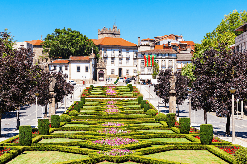 A view through a long narrow garden in Guimaraes towards stately white buildings with terracotta roofs. Trees line the garden.