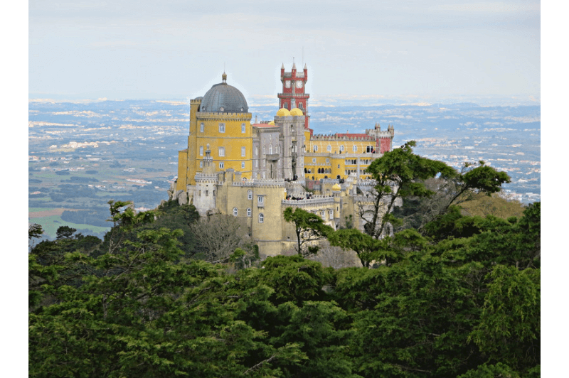 Pena palace in Sintra Portugal sits atop a lush green mountain. The castle is an ecclectic mix of towers, domes, and bright colours including mustard and red.