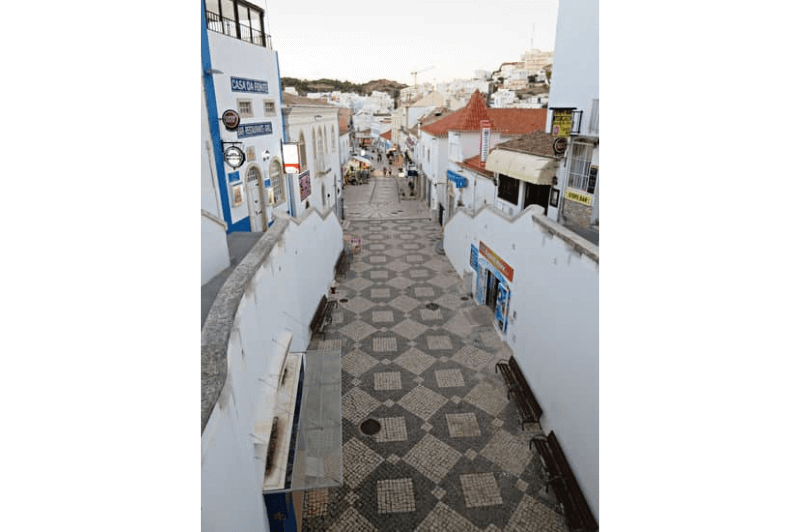 A patterned black and white cobbled street in Albufeira's old town. Whitewashed buildings with red roofs and blue accents line either side of the narrow street.
