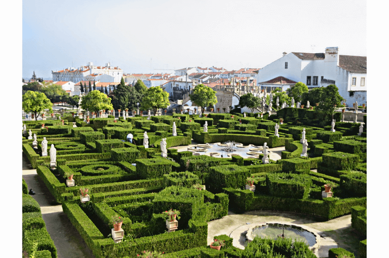 Bishop's Palace Gardens in Castello Branco. A baroque garden with structured hedges and statues throughout. A fountain is in the foreground and the whitewashed city is visible behind.