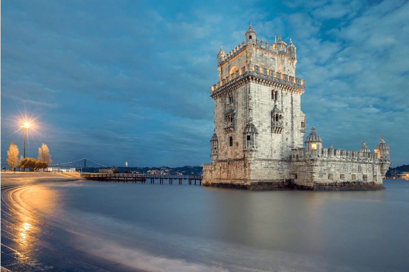 The gothic watch tower Belem rises out of a timelapse of the ocean. Bubbly clouds fill the evening sky and a light is on in the distance.