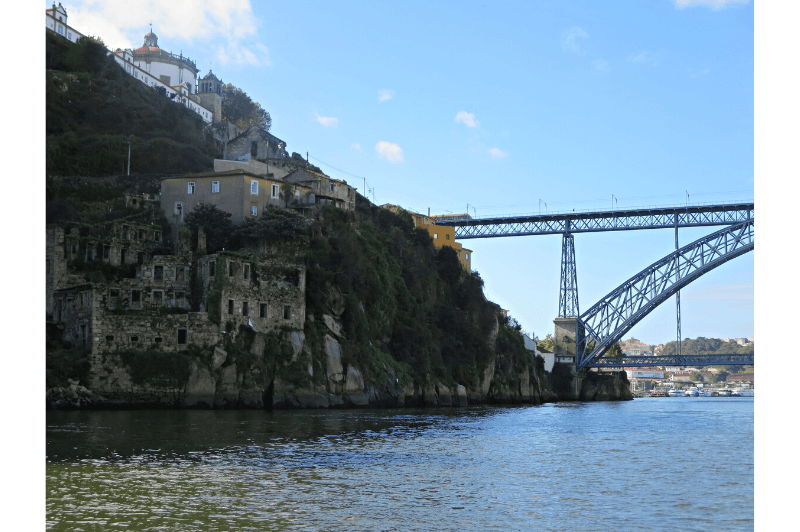 A photo from the Douro river in Porto Portugal looking towards the ruins of a large stone building which is built into the hillside of the rocky shoreline. A steel bridge with eiffel tower like construction reaches over the river to a hilltop behind the houses