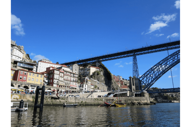 A photo from Porto Portugal on the Douro river looking towards the tall skinny row houses above the rocky shoreline. A steel bridge with eiffel tower like construction reaches over the river to a hilltop above the houses.