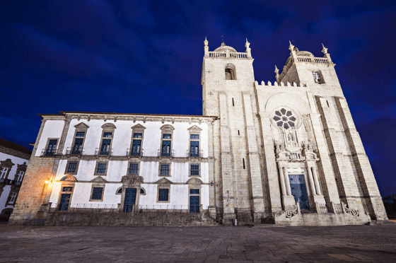A night shot at Porto Cathedral: A tall stone building reminiscent of Notre Dame adjoins a simple white Portuguese style rectangle building.