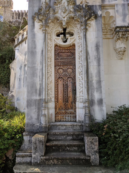 A wooden side door and stone steps at the gothic Manueline chapel on the estate of Quinta da Regaleira. The sun shines on the surrounding greenery.