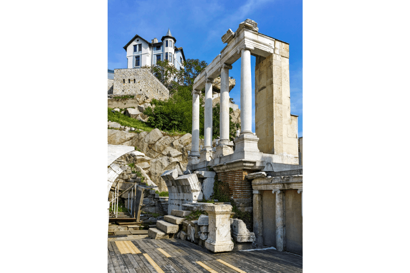 A side angle of the Plovdiv Amphitheatre with a mansion on a rocky hilltop in the background