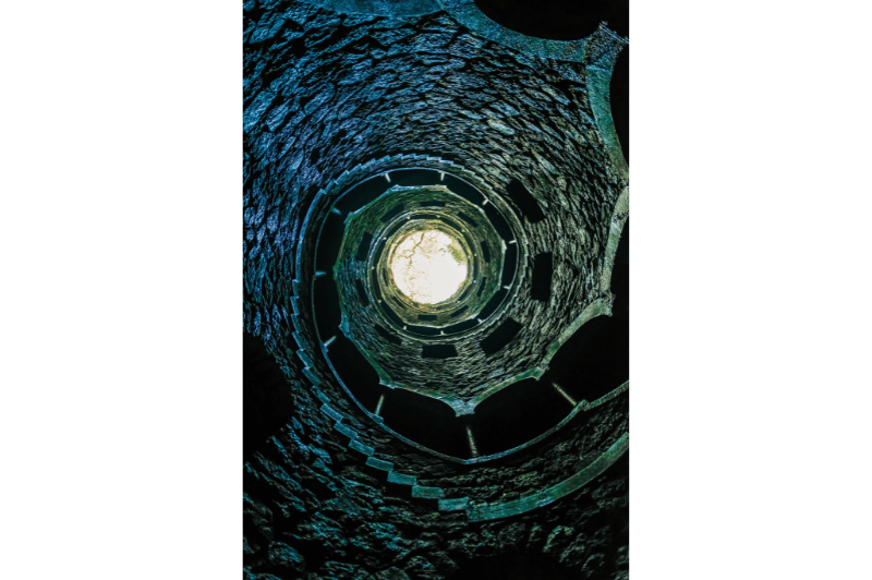 A dark shot of the Initiation well at Quinta da Regaleira looking up from the bottom to a circle of daylight at the top of the buried spiral staircase.