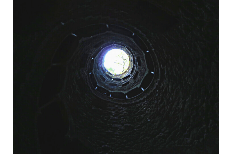 A dark shot of the Initiation well at Quinta da Regaleira looking up from the bottom to a circle of daylight at the top of the buried spiral staircase.