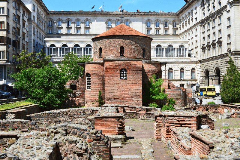 An ancient brick domed roof church in front of an 18th century building with 3rd century ruins in the foreground from early Serdica. St. George Church Rotunda Sofia near Vitosha Boulevard.