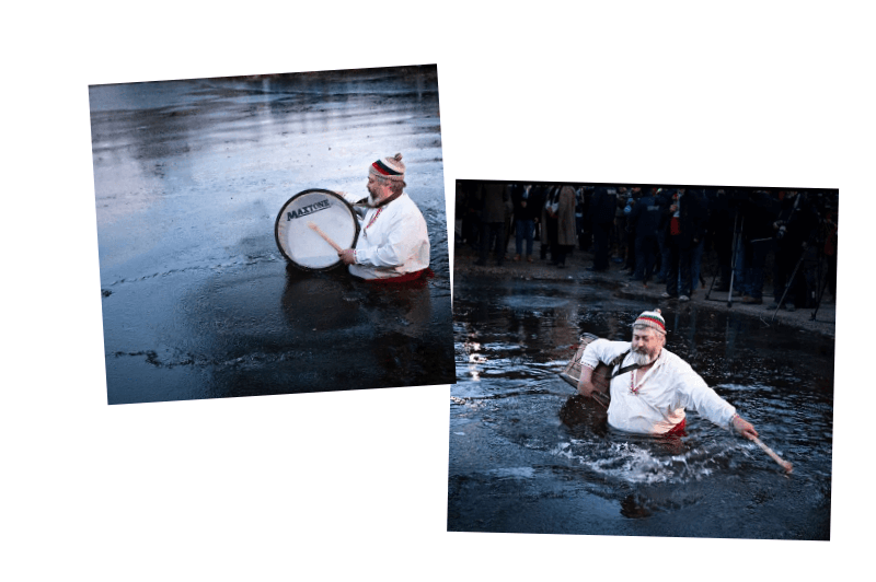 A portly older man in a Bulgarian folk shirt plays a drum in the icy chest deep water of a river for Iordanov Den