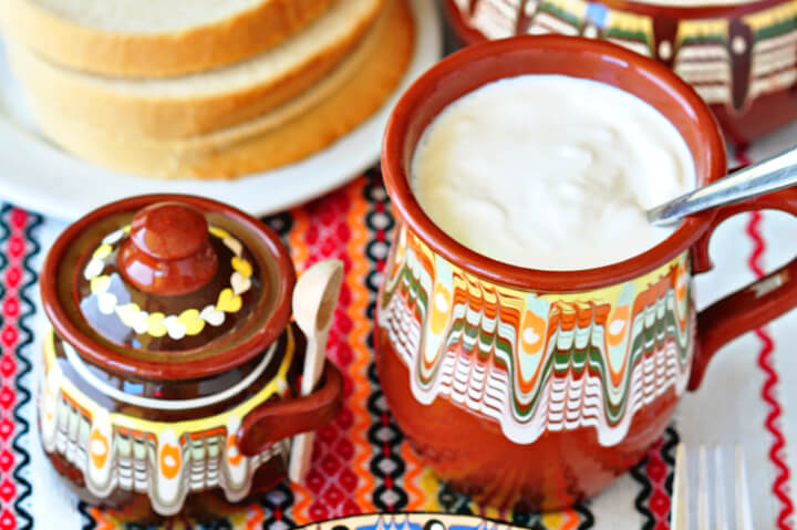 Traditional Bulgarian Clay pottery - rusty colored with a pattern of white, orange, green and yellow. One is a mug with handle that is full of yogurt with a spoon in it. The other is a small pot with a lid. Background is a plate of toast and the items are on a multi-colored folk table cloth.