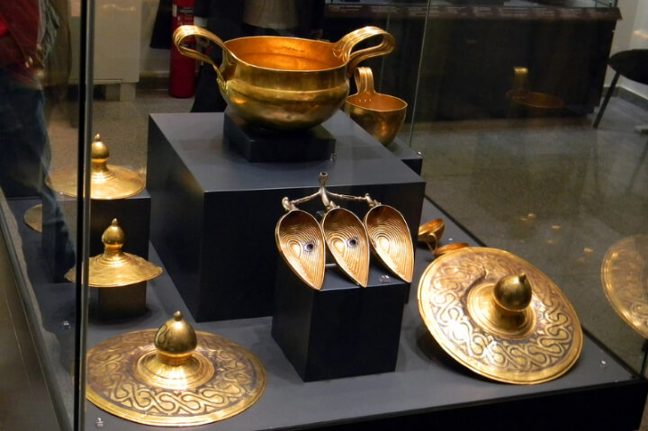 Six gold items of Thracian treasure in a glass museum case. A two handled bowl is on a block above the rest of the objects, and is the bowl a Bulgarian farmer fed his pigs out of.