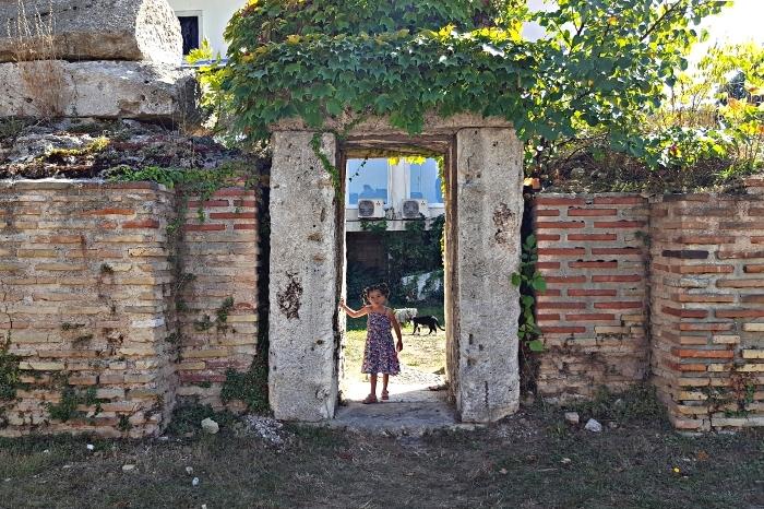 A small girl poses in an ancient ROman doorway, left standing in a grassy area in Varna. Vines grow over the doorway and a black and white cat walks behind her.