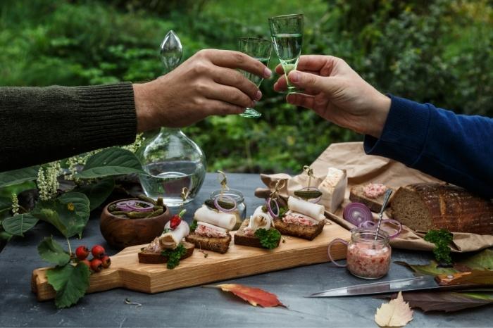 Two hands cheers with mastika in small shot glasses over a charcuterie picnic.