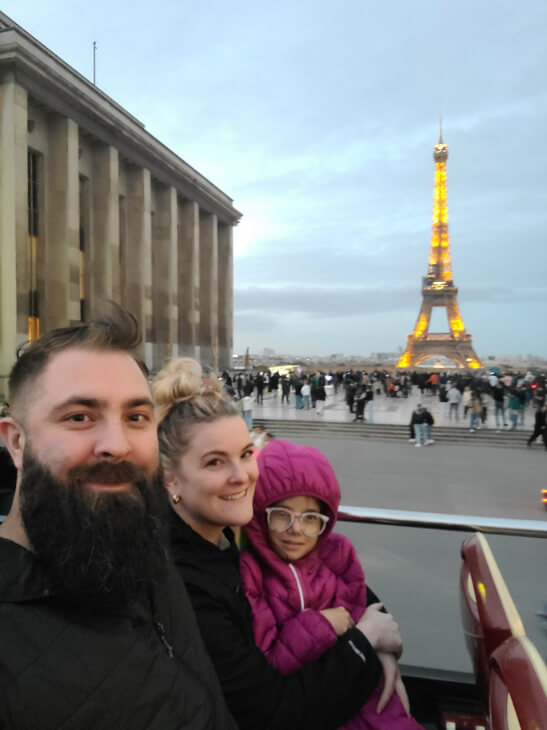A family smiles in the top of a double decker tour bus in front of the eiffel tower. Our first stop during our gap year!