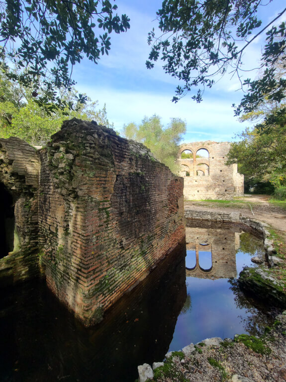 Fountain of the Nymphs at Butrint National Park. A ruined stone building stands in a pool of water. Reflected in the pool is a stone archway from the distant Great Basilica.