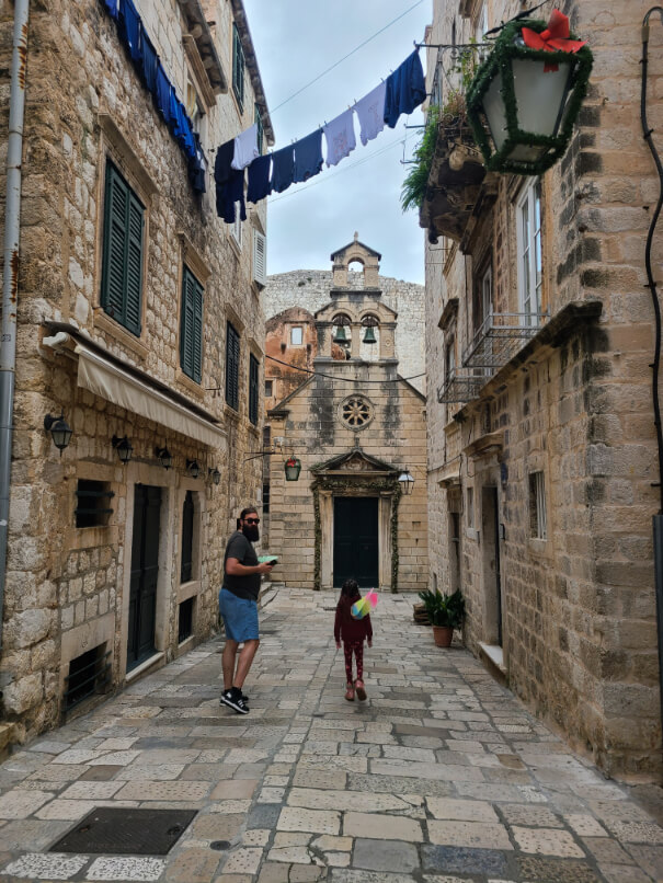 Man with sunglasses turns to the camera in a street in Dubrovnik where he stands beside a girl in fairy wings. The lanterns are decorated for Christmas, and so is a church in the background. All blue laundry hangs on a line overhead.