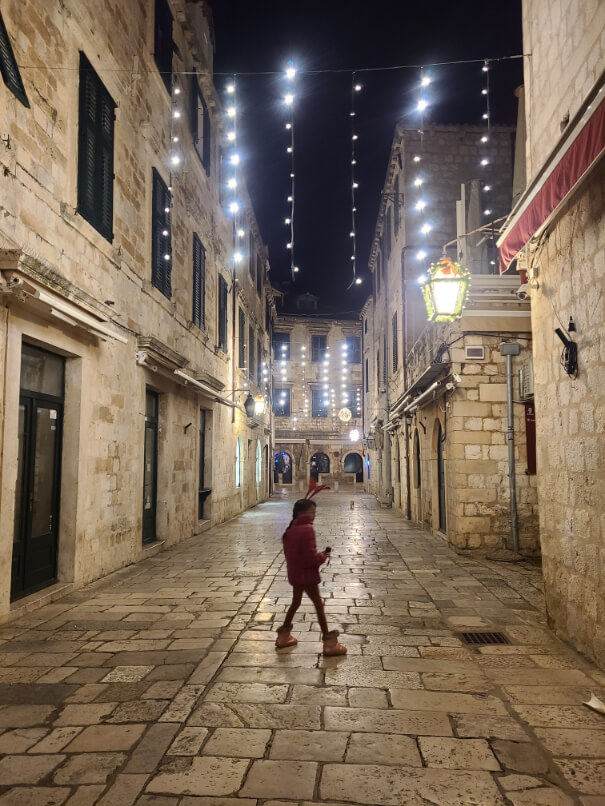 Child crosses the street under some Christmas Lights in Dubrovnik at night. She is wearing a pink coat and reindeer antlers.